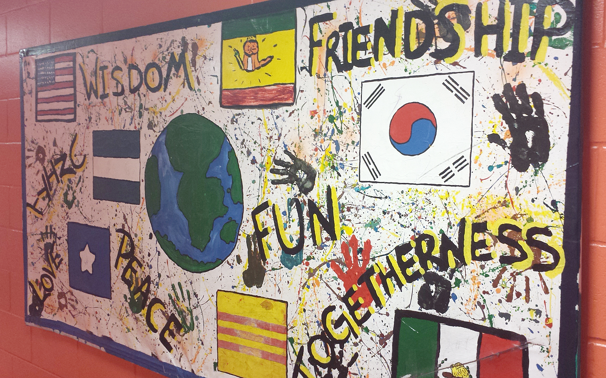 Painting hanging on a red wall with the earth in the middle, flags or various countries, handprints, and the words, "wisdom", "friendship", togetherness", "fun", "peace", "love", and a word that cannot be made out.